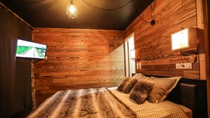 Ein Zimmer mit Holzwänden, einem Bett mit gemustertem Überwurf, zwei Kissen, einem Wand-Fernseher und einer Pendelleuchte.