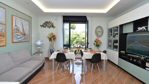 A modern living room with a dining area, a sofa, a TV, and a large window with a view of greenery.