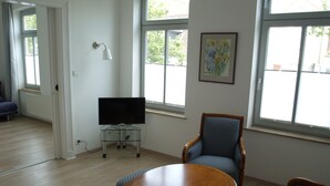 Una sala de estar con una mesa de madera, dos sillas azules con motivos y un televisor sobre un pedestal de vidrio, además de un cuadro en la pared.