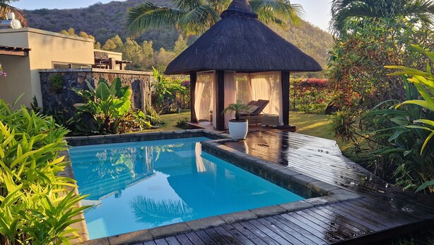 Une piscine dotée d’une terrasse en bois, entourée d’une végétation luxuriante et bordée d’une structure à toit de chaume.