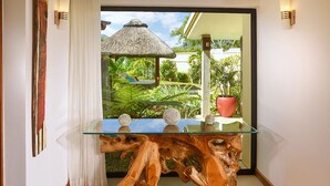 Une table dont le plateau est en verre et dont le piétement est en bois, un rideau blanc et une vue sur un jardin tropical agrémenté d’une structure à toit de chaume.