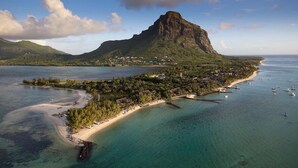 Une vue aérienne d’une zone côtière, avec une montagne imposante, des eaux cristallines et une promenade aménagée le long de la plage, bordée de bâtiments et de bateaux.