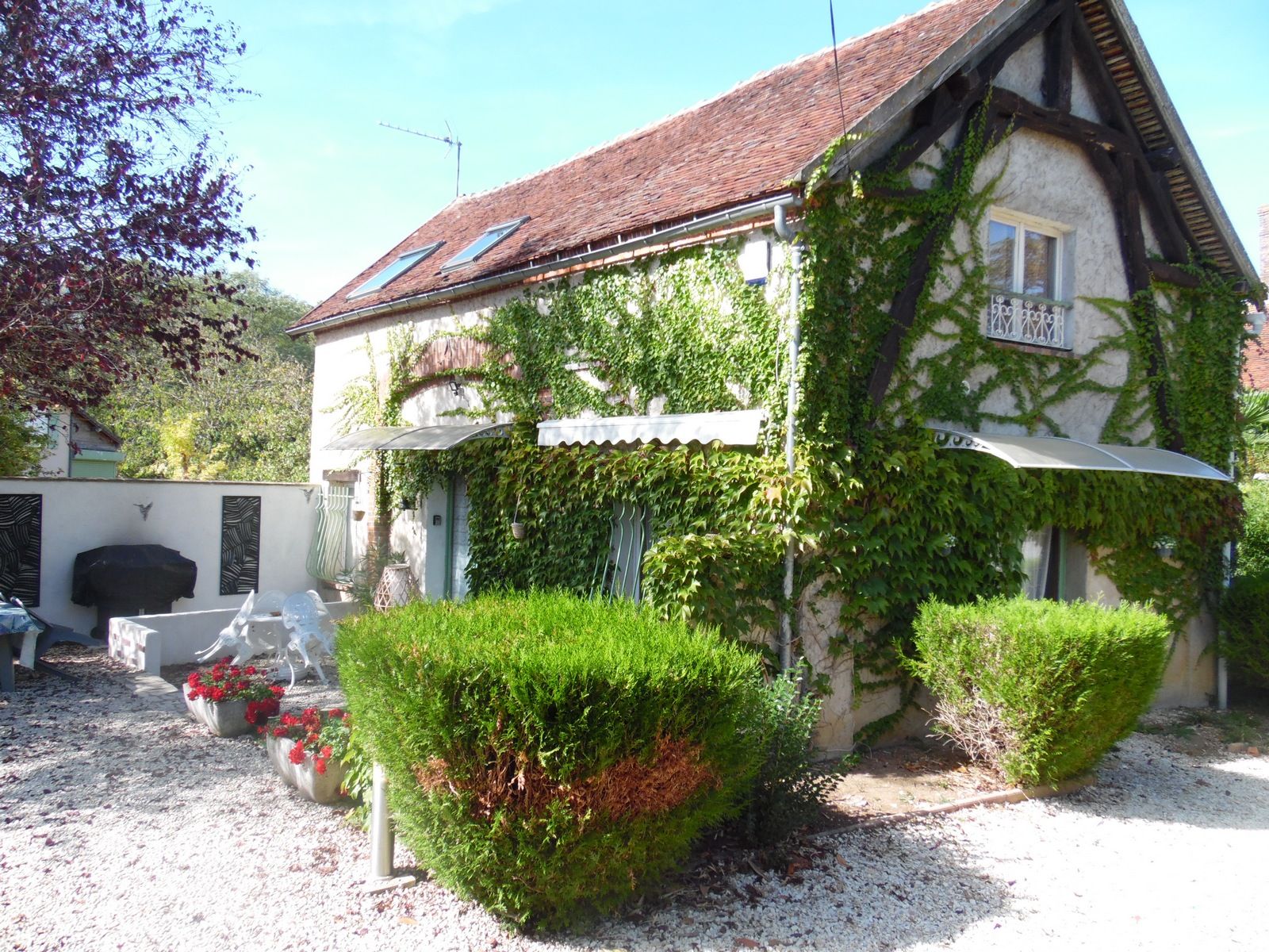 Un bâtiment aux murs recouverts de lierre, doté d’un toit en tuiles et d’un petit jardin agrémenté d’arbustes taillés et de plantes en pot.