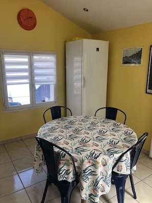 Un coin repas avec une table ronde recouverte d’une nappe à motifs floraux, entourée de quatre chaises. Un réfrigérateur blanc est adossé au mur, et une horloge est fixée au mur au-dessus de la fenêtre.