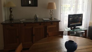 Ruokailutila, jossa on puinen pöytä, tuoleja, maljakko, televisio lasipöydällä ja ikkuna, jossa on verhot.