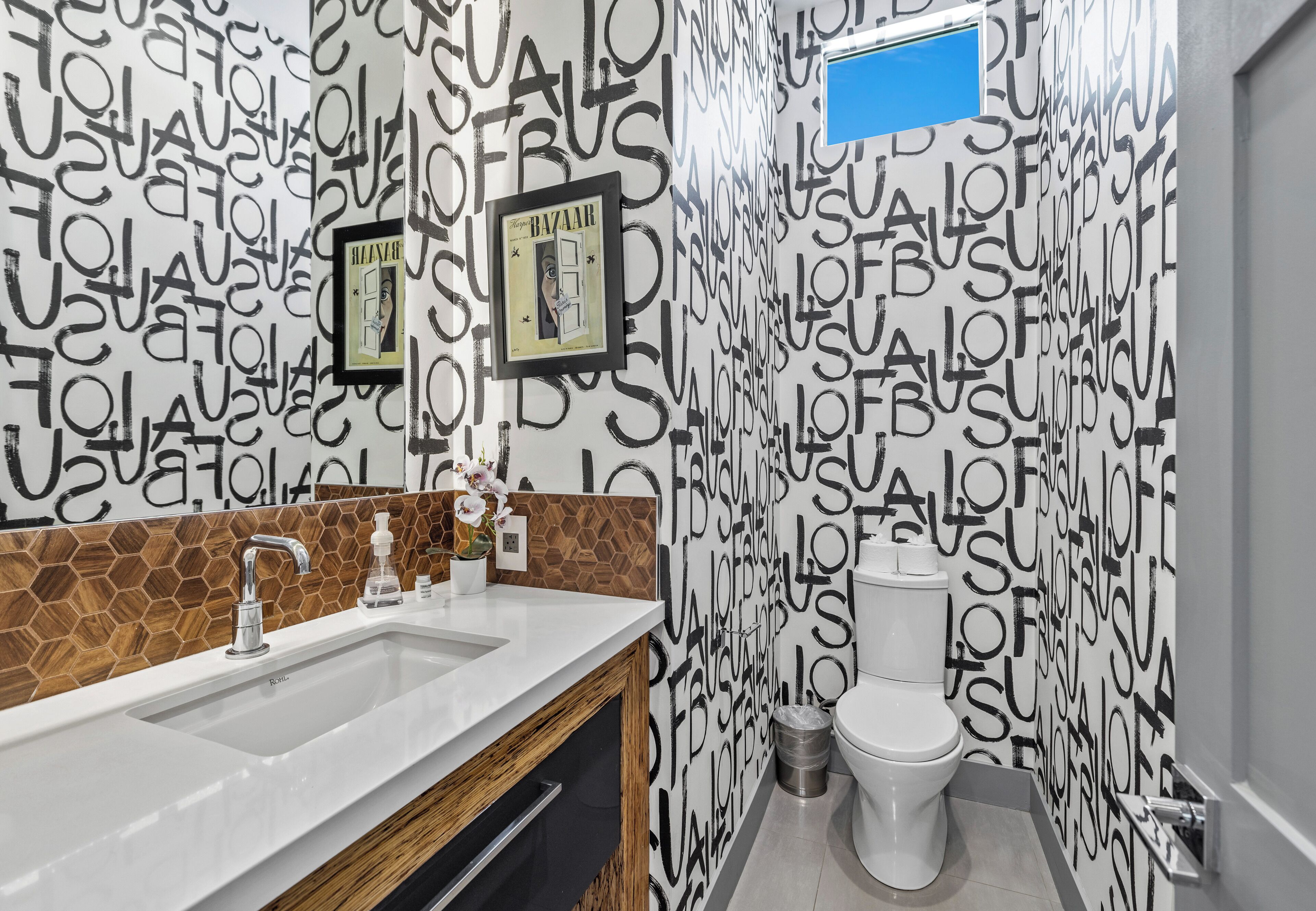 A bathroom with a white sink, a mirror, and a toilet. There is a framed picture on the wall and a trash can next to the toilet.