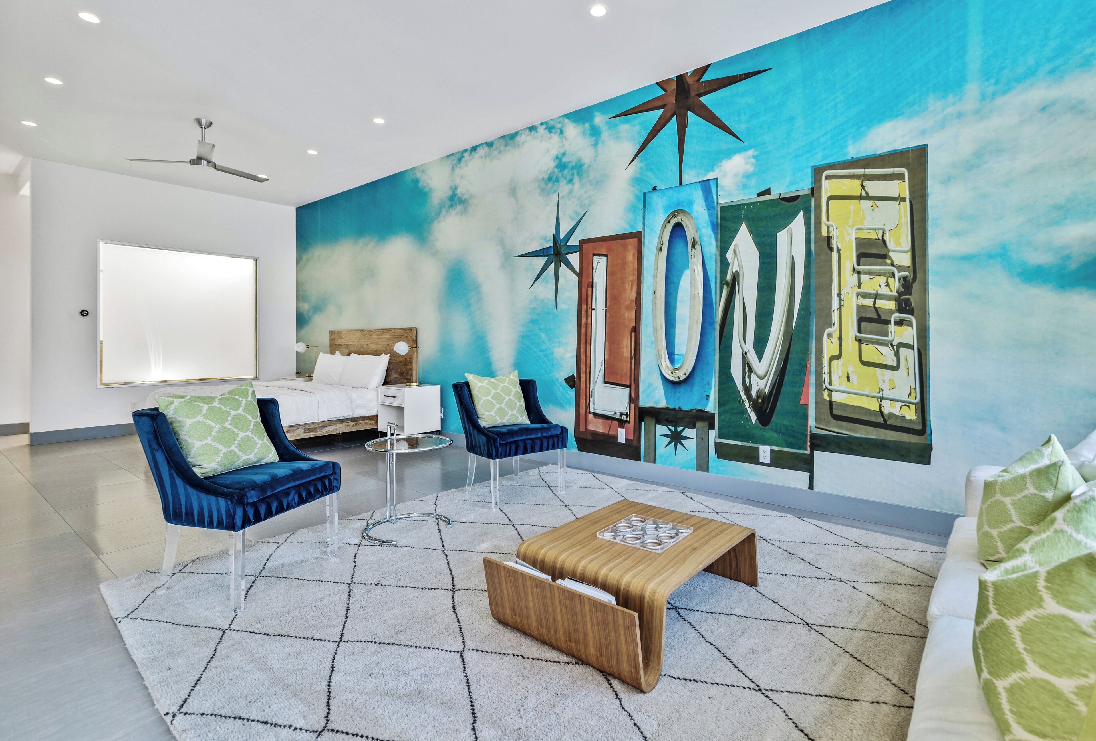 A modern hotel room with a large mural, a bed, two armchairs, a coffee table, and a ceiling fan.