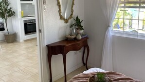 Een slaapkamer met een bed, nachtkastje met spiegel, raam met gordijnen en uitzicht op een keuken.