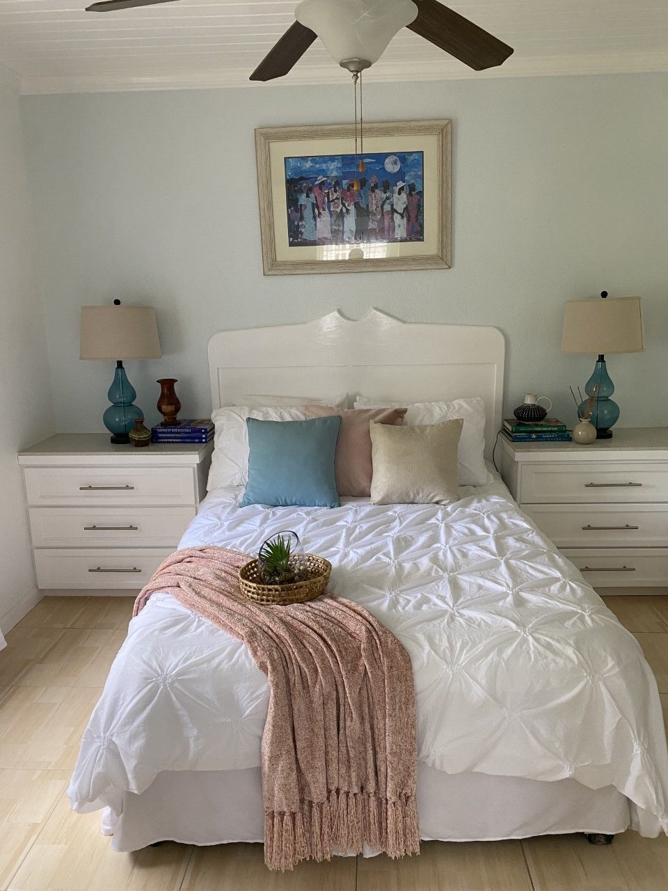 A bedroom with a white bed, two nightstands, a ceiling fan, a framed painting, and a small plant on the bed.
