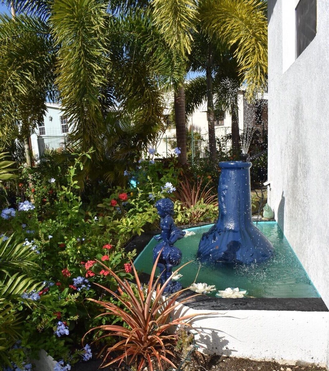 A garden with a fountain, various plants, and a white building in the background.