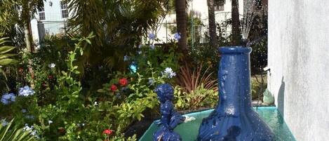 A garden with a fountain, various plants, and a white building in the background.