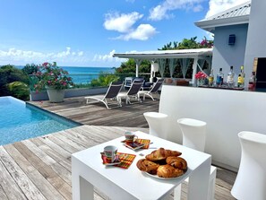 A poolside area with lounge chairs, a table with croissants and a cup, and a view of the ocean. - Exceptional villa with sea view, infinity pool -Sainte Anne-12 people (Sainte Anne)