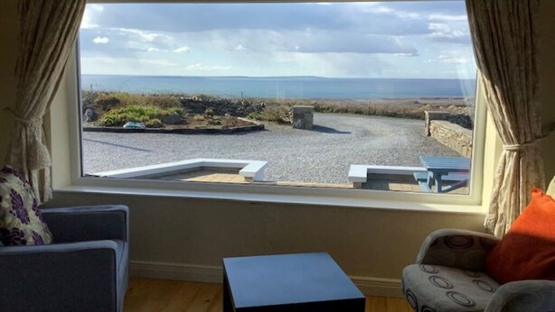 Una habitación con un ventanal grande con vistas a una playa y un paisaje rocoso. Hay dos sillas y una pequeña mesa dentro.