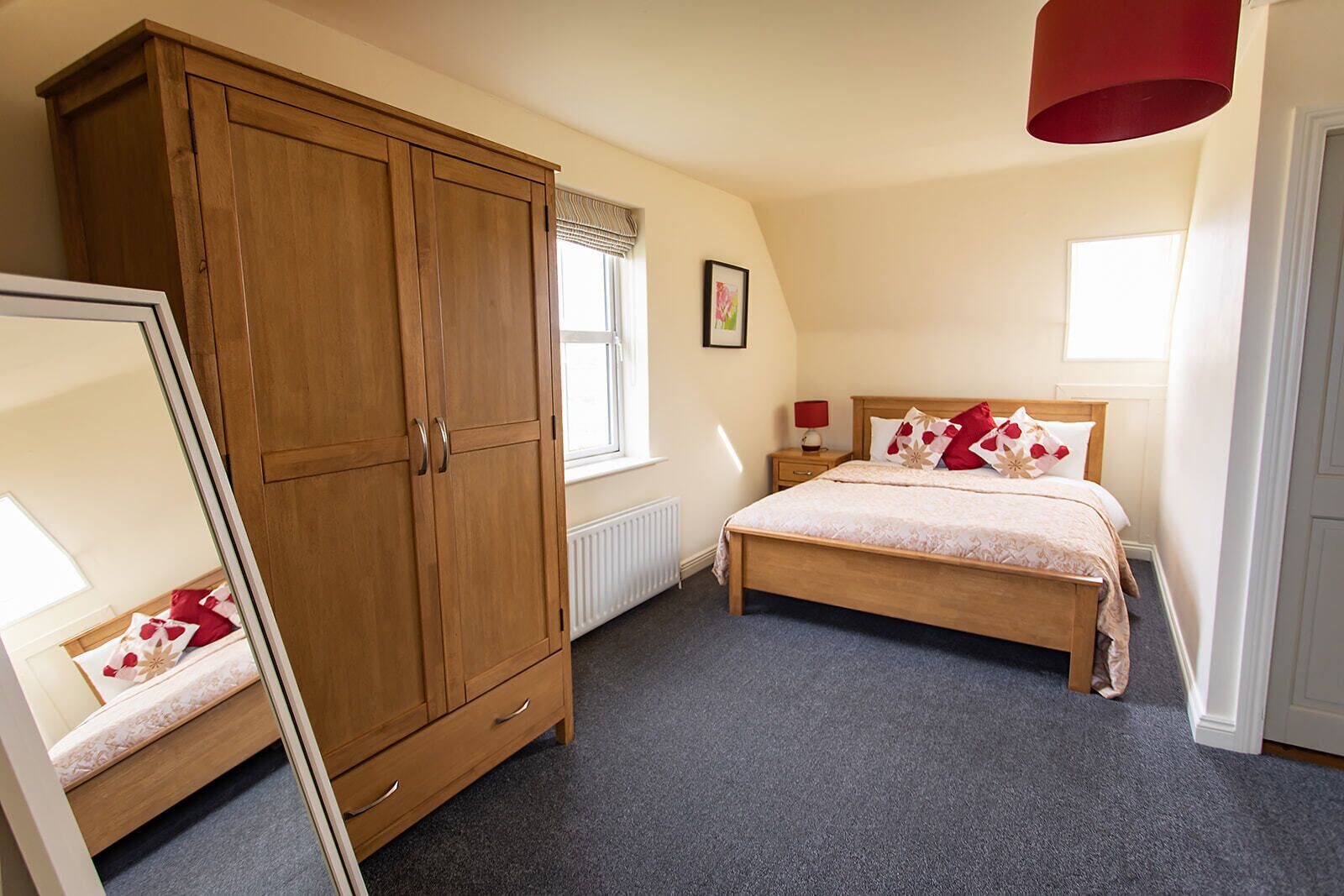A bedroom with a large wooden wardrobe, a bed with floral bedding, a bedside table with a lamp, and a window with blinds.