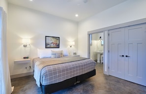 A bedroom with a bed, bedside lamps, a painting, a wardrobe, and a bathroom visible through an open door.