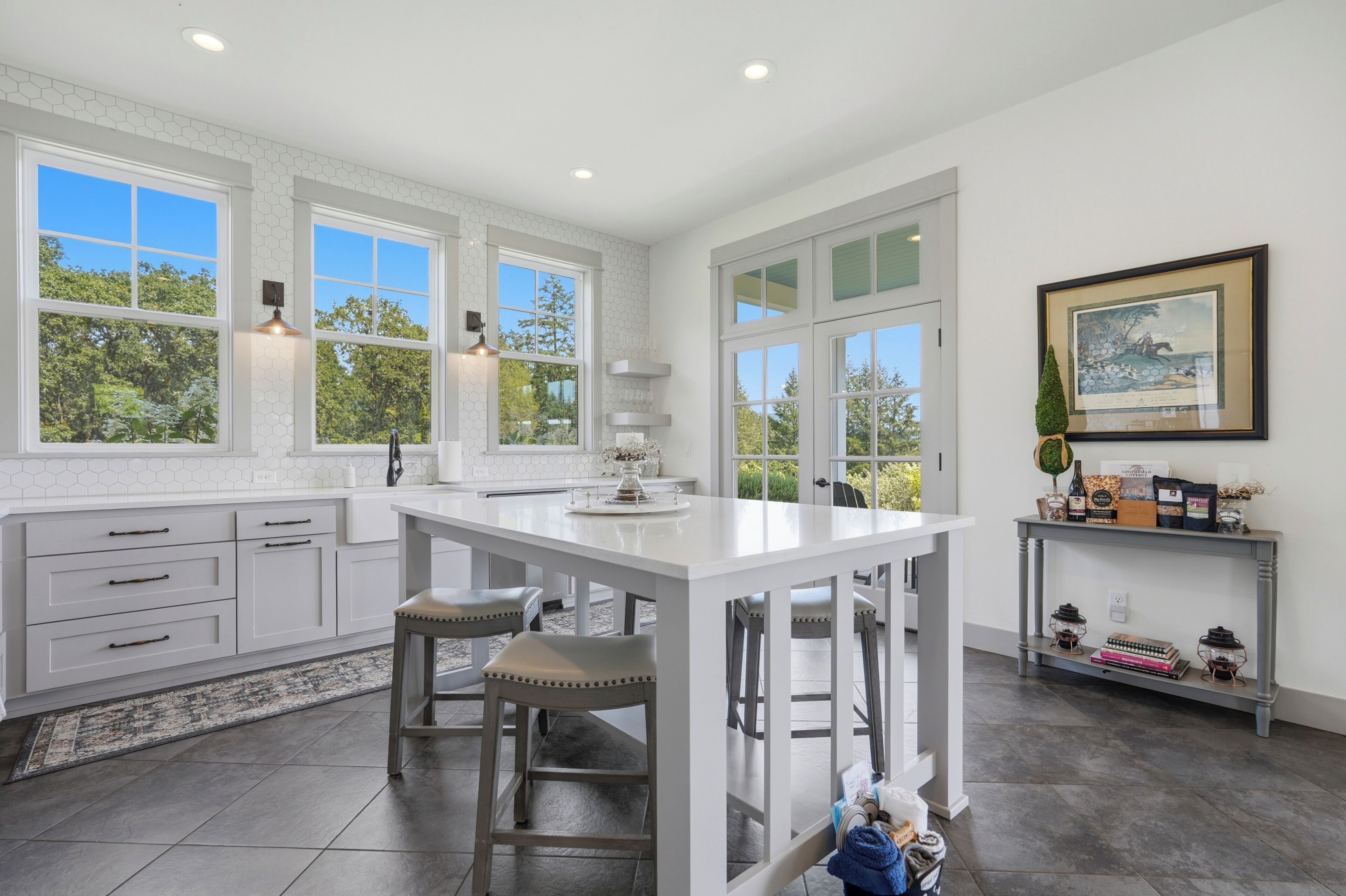 A modern kitchen with white cabinetry, a central island, and large windows offering views of greenery.