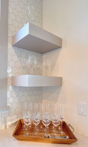A kitchen with three white shelves holding wine glasses and a wooden tray with more wine glasses and a corkscrew.