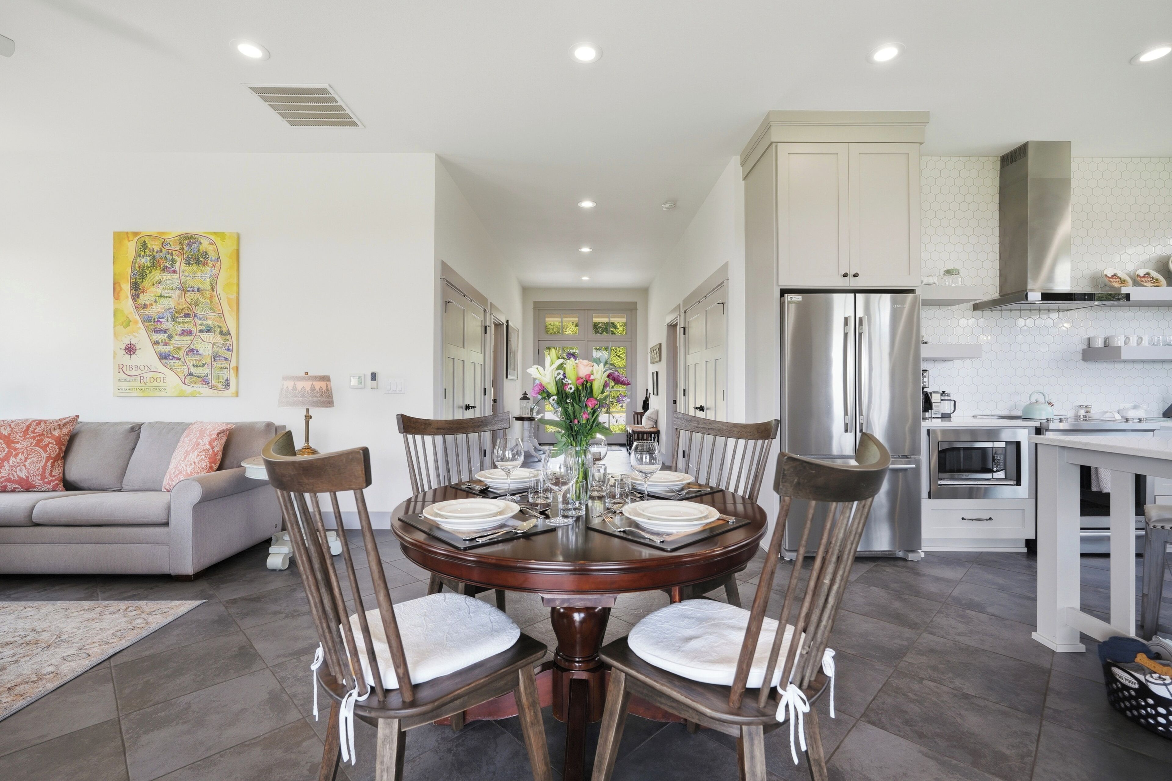 A modern kitchen with stainless steel appliances, a dining area with a round table and chairs, and a living room with a sofa and a decorative wall piece.