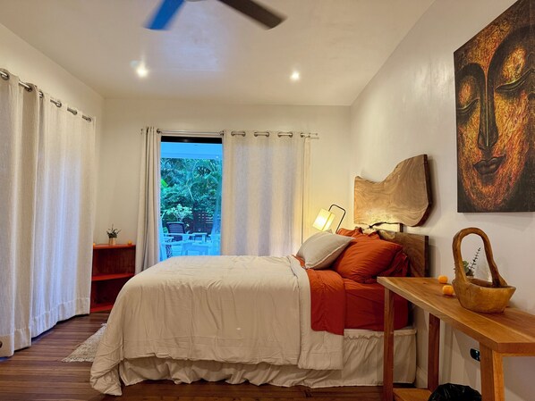 Un dormitorio con cama, mesita de noche, lámpara, un cuadro y vistas al exterior.
