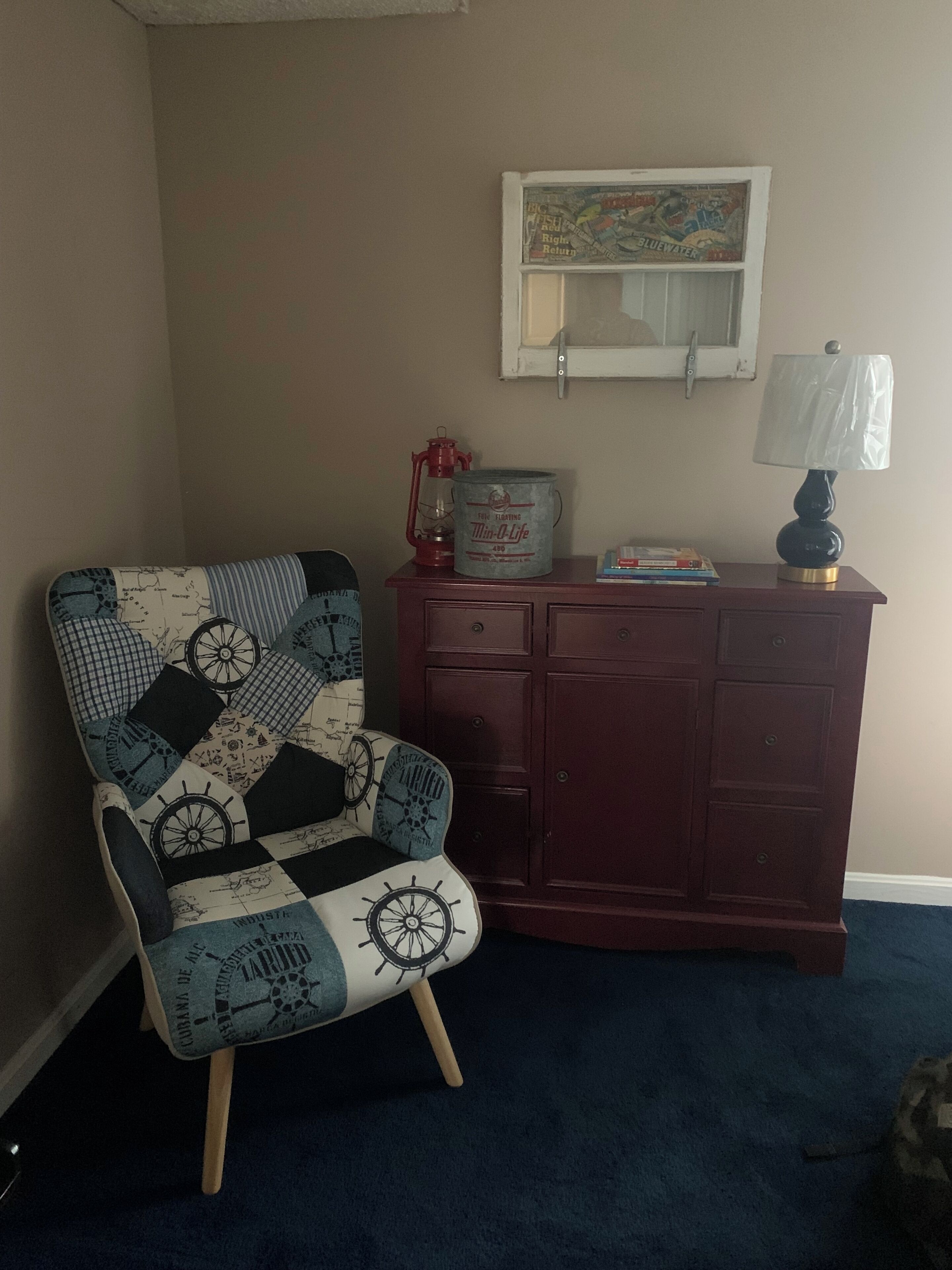 A room with a navy blue carpet, a navy blue armchair with a nautical theme, a wooden dresser, and a lamp.