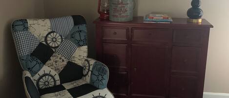 A room with a navy blue carpet, a navy blue armchair with a nautical theme, a wooden dresser, and a lamp.
