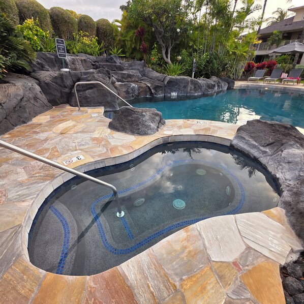 A hot tub with a tiled edge, surrounded by rocks and a pool.