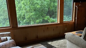 A cozy room with wooden walls and floors, large windows offering a forest view, a sofa, a chair, and a small table. - Simply Irresistible: Authentic Log Cabin (Creston)