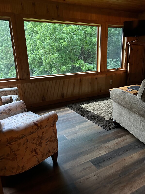 Een gezellige kamer met houten wanden en vloeren, grote ramen met uitzicht op een bos, een bank, een stoel en een kleine tafel.