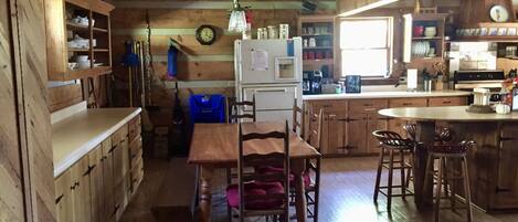 A wooden cabin interior with a dining table, chairs, a kitchen area with a refrigerator, and a clock on the wall.