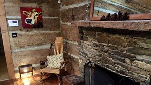 A cozy room with a stone fireplace, a wooden chair with a cushion, and a painting of a cow on the wall. - Simply Irresistible: Authentic Log Cabin (Creston)