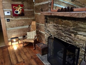 A cozy room with a stone fireplace, a wooden chair with a cushion, and a painting of a cow on the wall. - Simply Irresistible: Authentic Log Cabin (Creston)