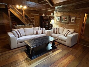 A rustic living room with a sectional sofa, a coffee table, wooden flooring, exposed beams, and a staircase.