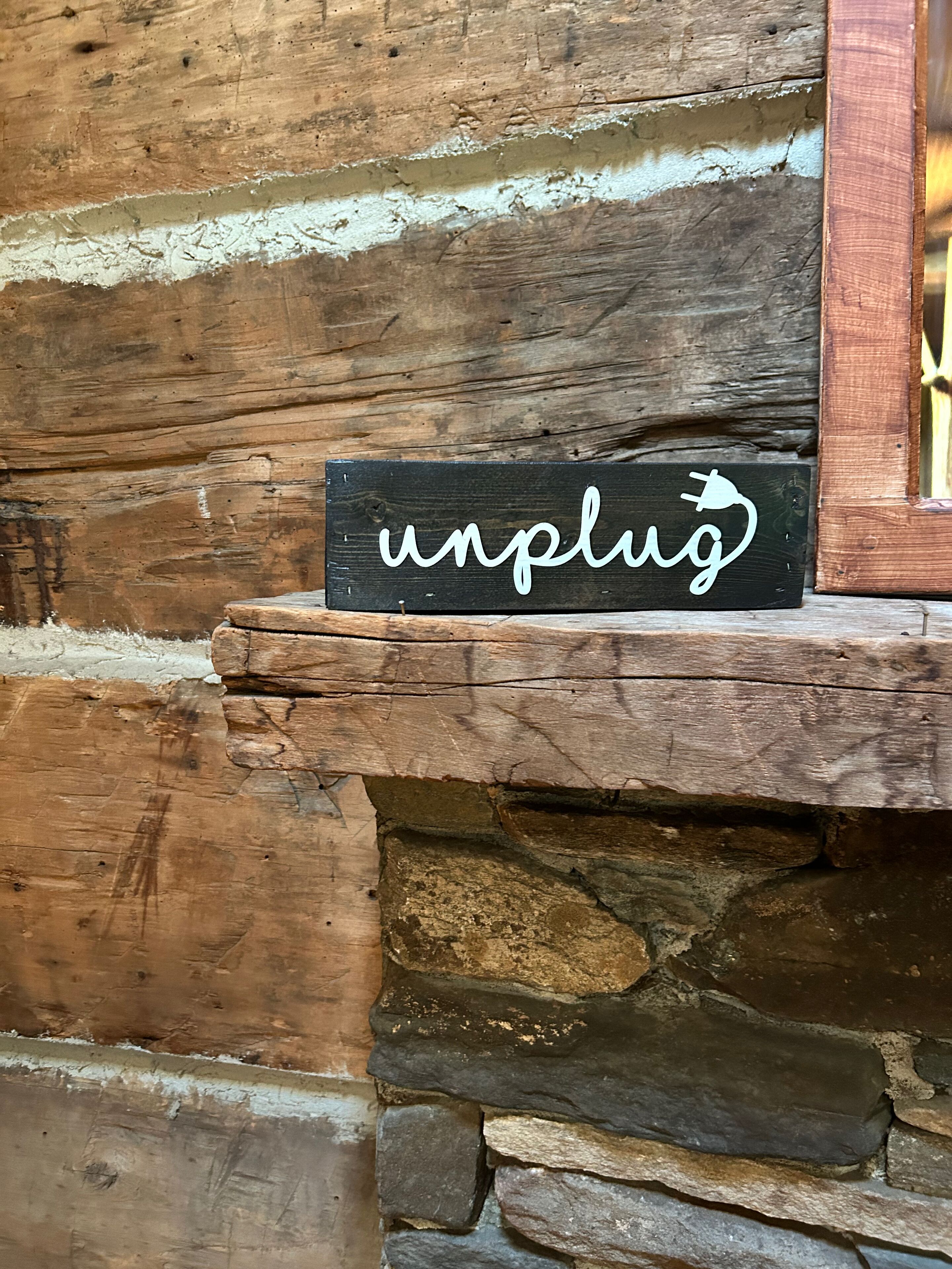 A rustic wooden sign with the word "unplug" and a plug icon, placed on a stone ledge against a wooden wall.