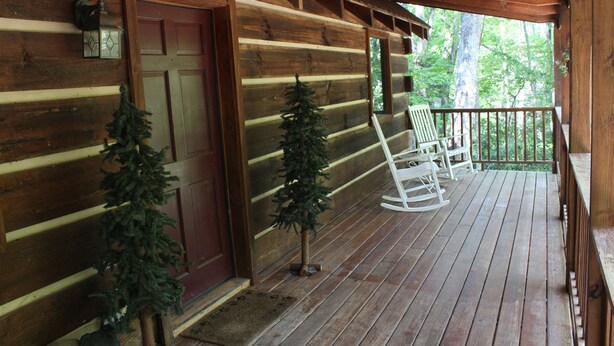 Une cabane en bois avec un porche, deux petits sapins de Noël et une chaise à bascule.