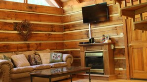 L’intérieur d’une cabane en rondins, avec une cheminée, un canapé, un téléviseur fixé au mur et un plafond en bois.