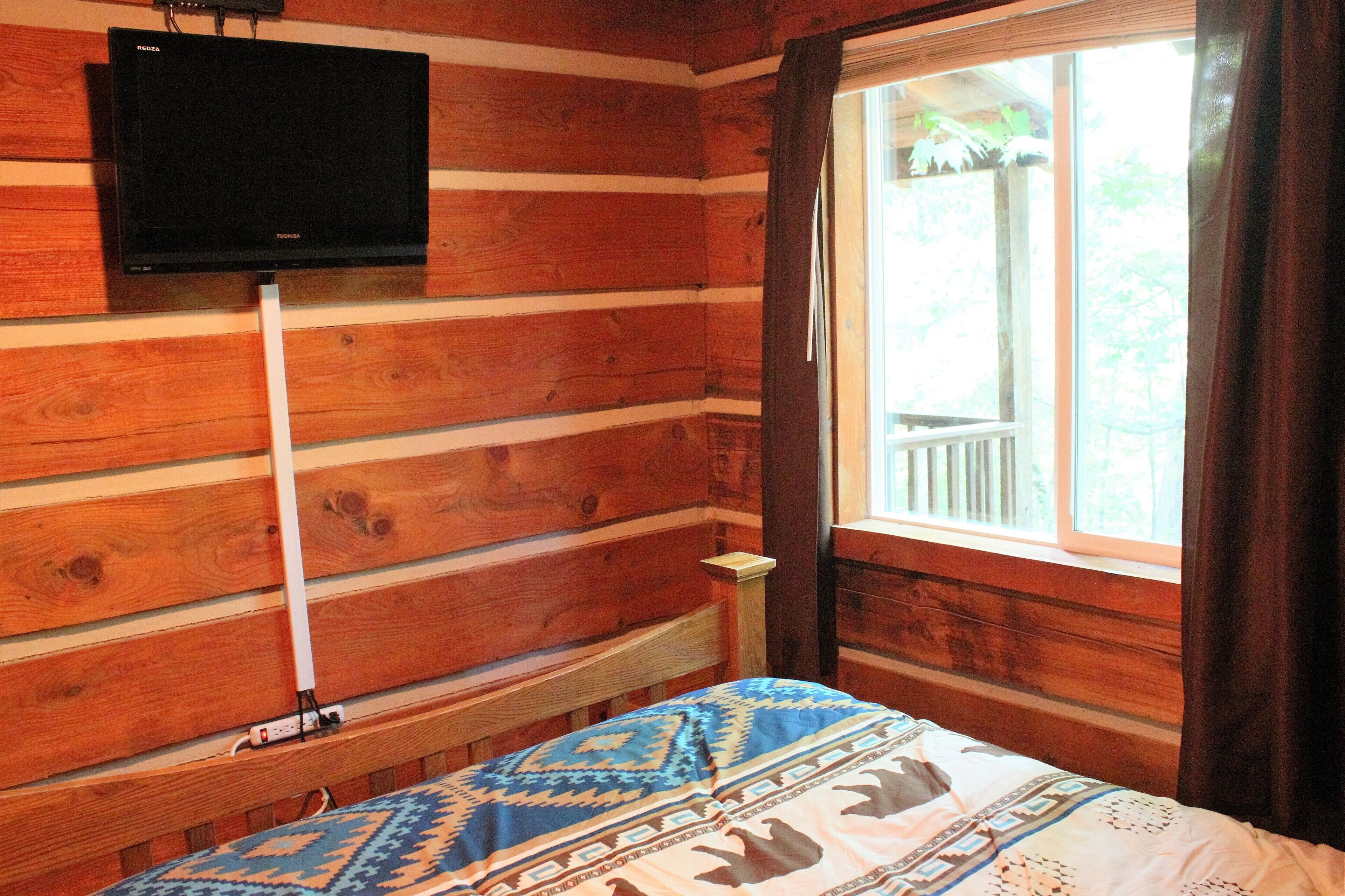 A wooden cabin room with a bed, a mounted TV, and a window with curtains.