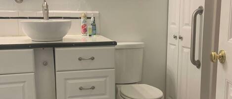 Une salle de bain avec un lavabo blanc, des toilettes et une fenêtre située au-dessus des toilettes. Il y a trois tiroirs sous le lavabo et un tapis noir sur le sol.