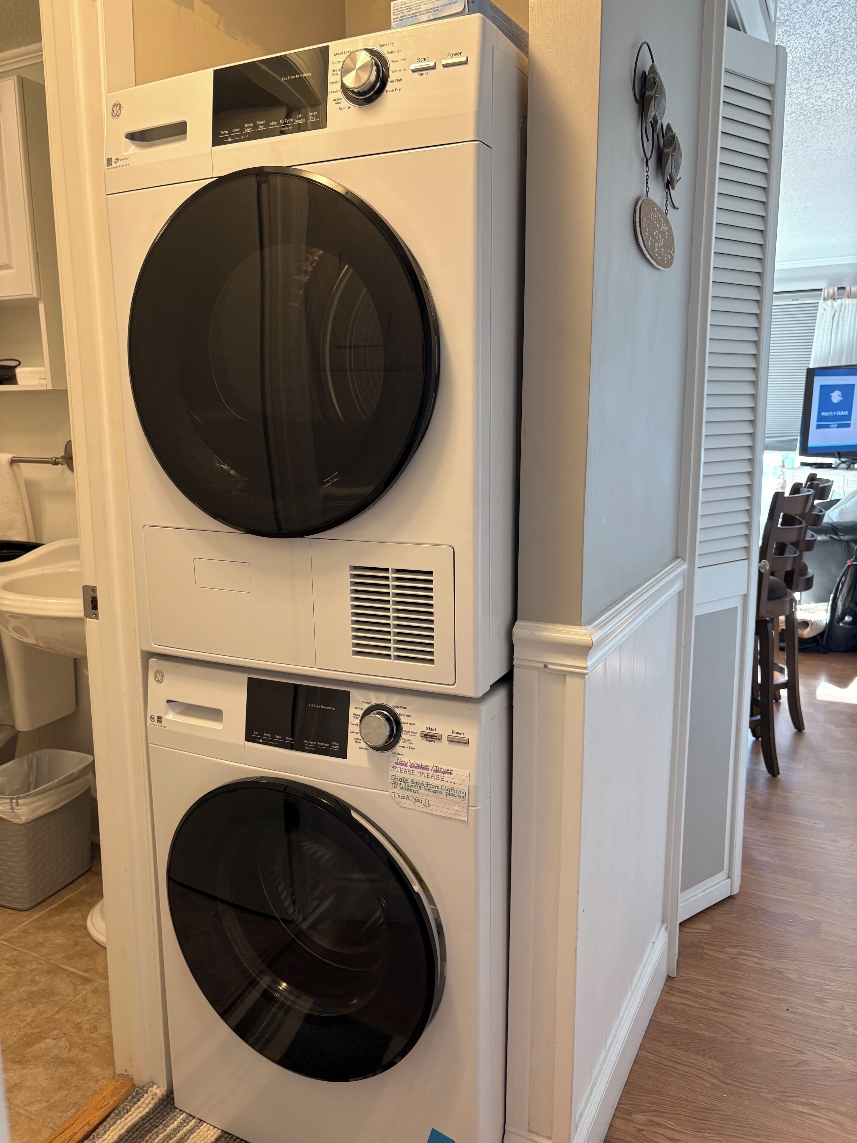 A stacked washer and dryer unit in a room with a wooden floor and a window with blinds.