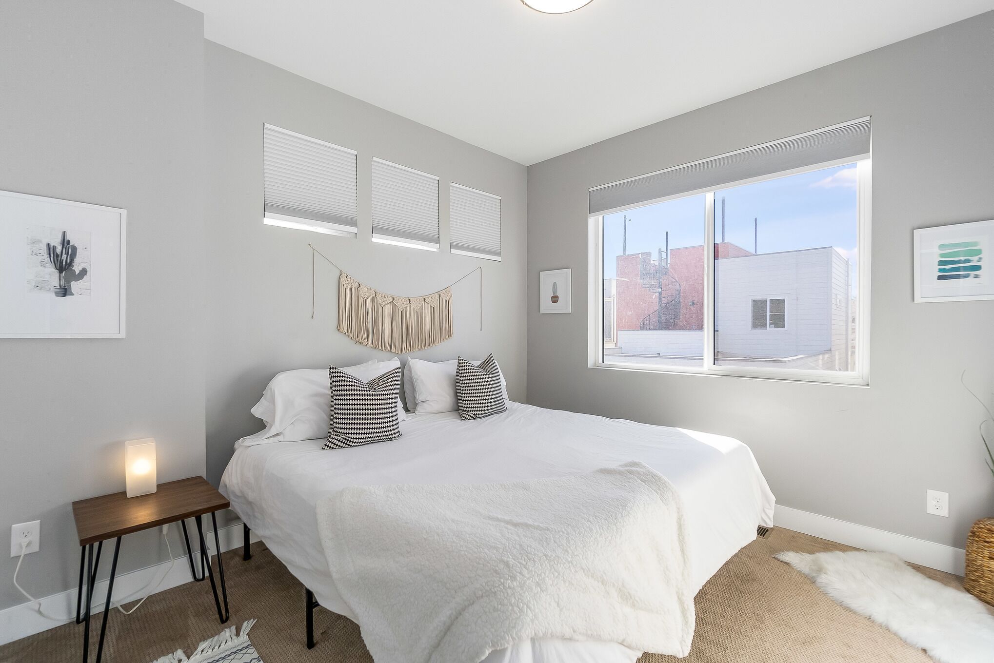 A bedroom with a large bed, a bedside table with a lamp, a window with blinds, and a view of buildings outside.