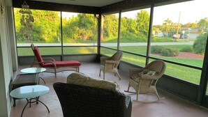 A covered outdoor seating area with wicker furniture and a glass table, overlooking a well-maintained lawn and distant buildings.