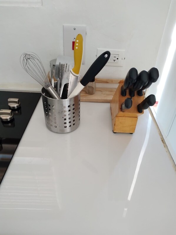 Una encimera de cocina con un portalatazajes que contiene un batidor, espátulas y un cuchillo. Hay una tabla de cortar de madera y un bloque para cuchillos con varios cuchillos.