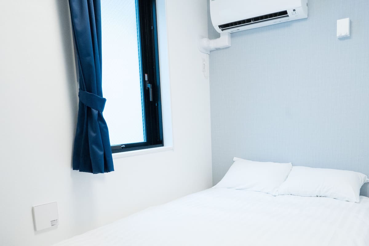 A single bed with white bedding, a window with blue curtains, and a wall-mounted air conditioning unit.
