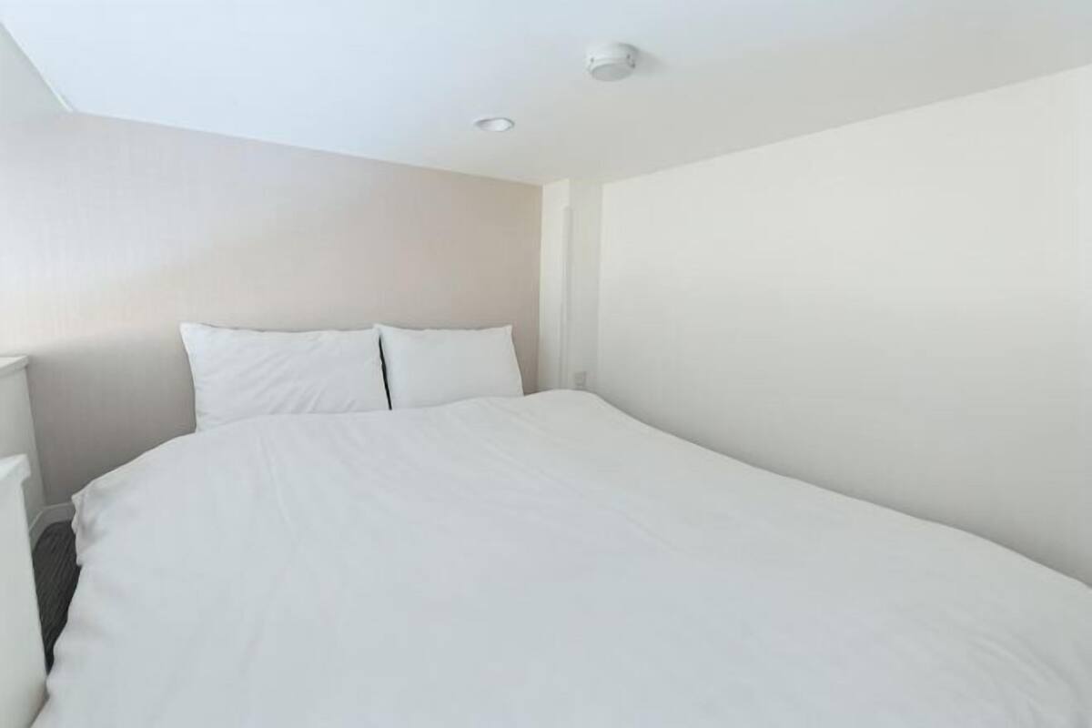 A single bed with white bedding in a room with plain walls and a ceiling light.