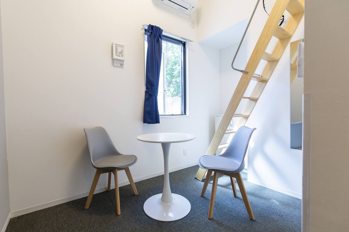 A room with a round table and two chairs, a window with a curtain, a wooden ladder, and a small picture frame on the wall.