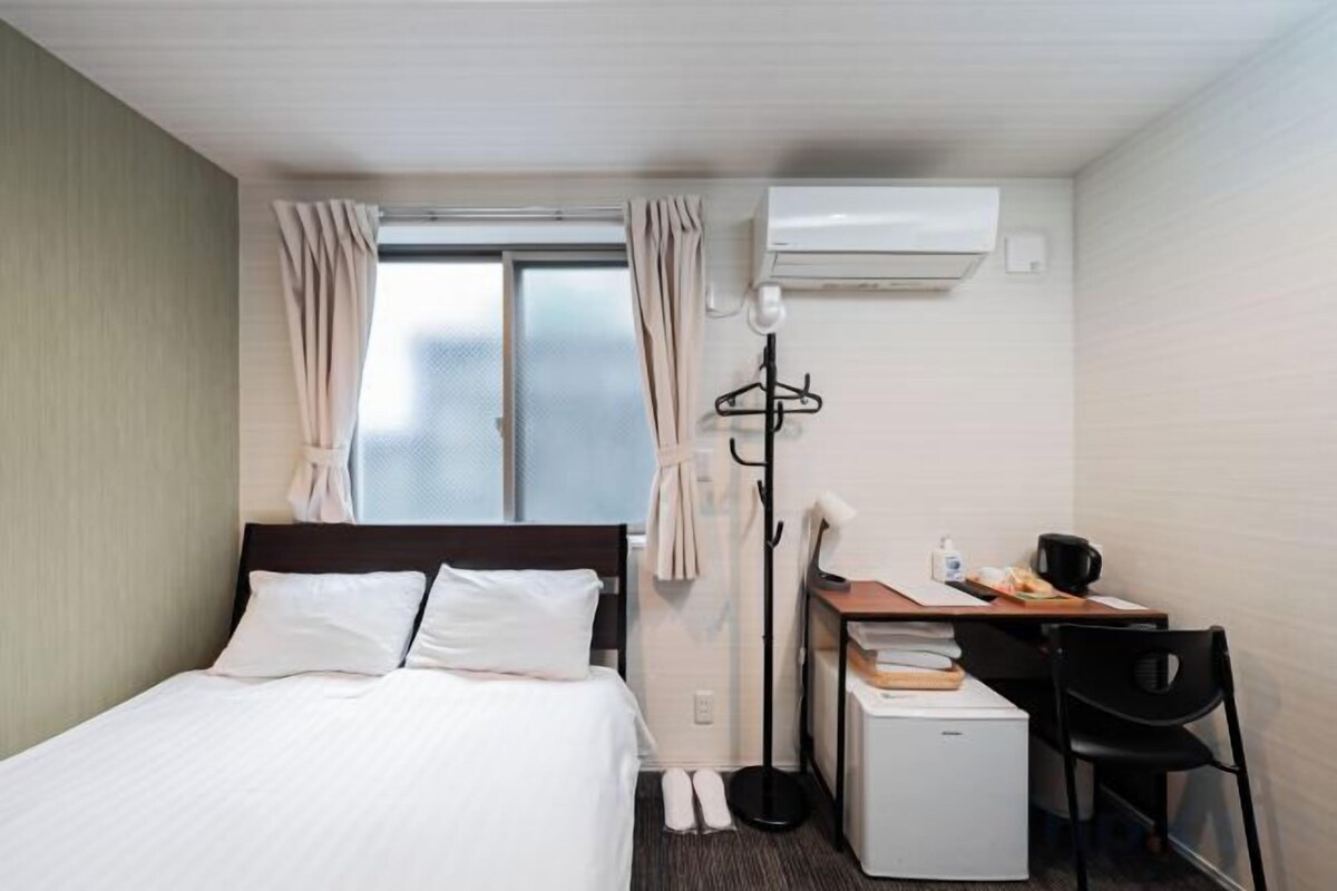 A hotel room with a bed, a desk, a chair, a small refrigerator, and an air conditioning unit.