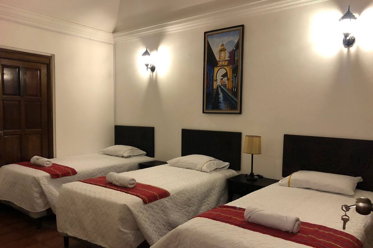 Una habitación de hotel con tres camas individuales, cada una con ropa blanca y detalles rojos, un cuadro de un puente en la pared y una puerta de madera.