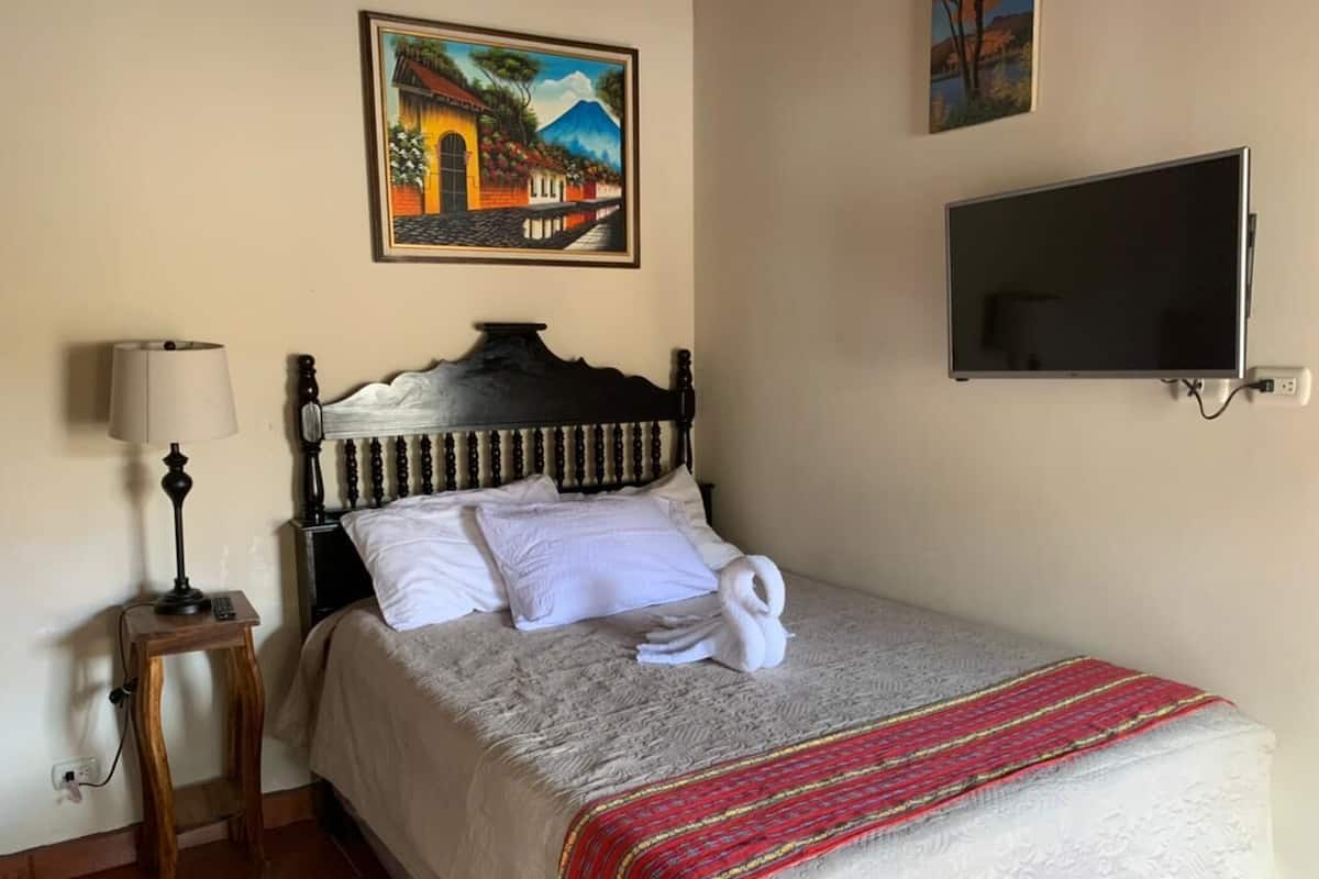 Una cama bien tendida con una colcha blanca y una manta a rayas dorada y roja, una mesita de noche de madera con una lámpara, un cuadro de un edificio en la pared y un televisor de pantalla plana montado sobre la cama.