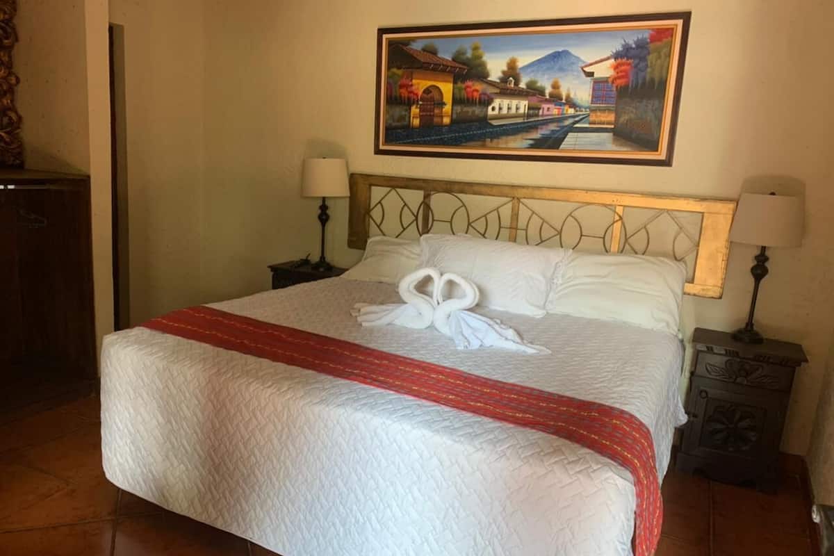 Una cama bien hecha con una colcha roja y blanca, dos almohadas blancas y un diseño decorativo de cisne en la colcha. Hay un cuadro de un paisaje en la pared sobre la cama.