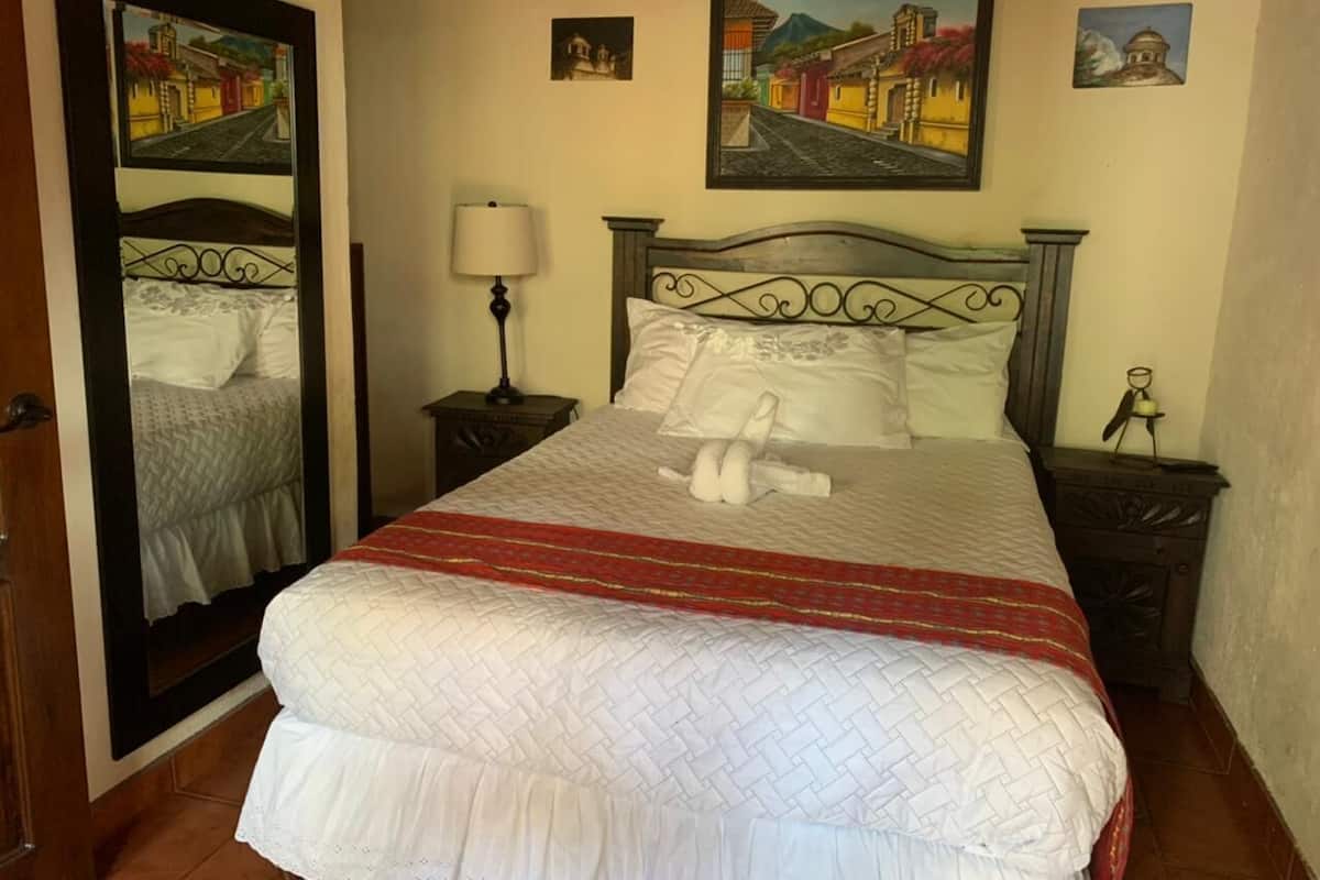Una cama bien hecha con colcha blanca y manta roja, cabecera de madera, lámpara de noche y una mesita con lámpara.