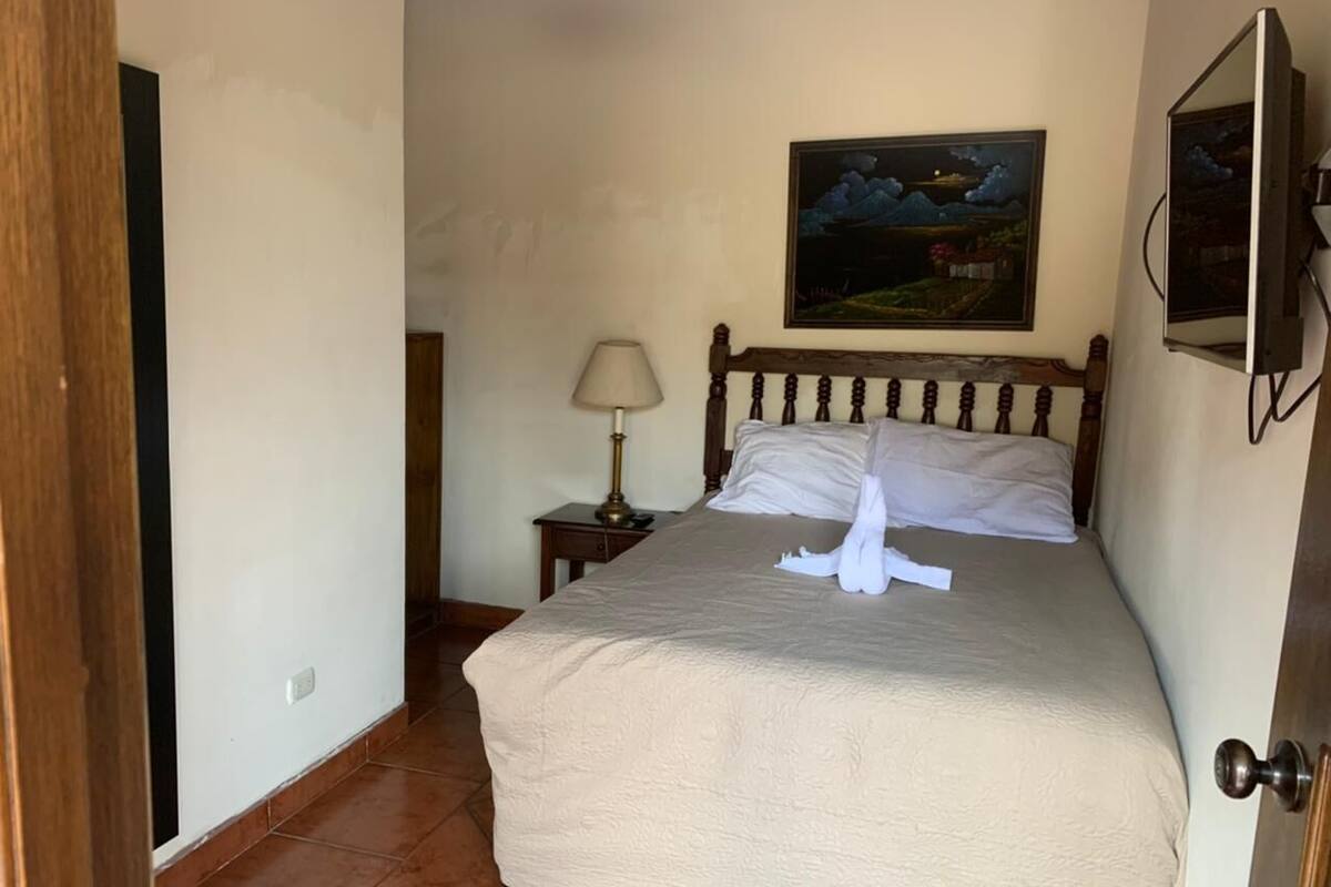 Una cama bien hecha con un edredón color beige y almohadas blancas, una lámpara de noche y un cuadro de un paisaje en la pared.